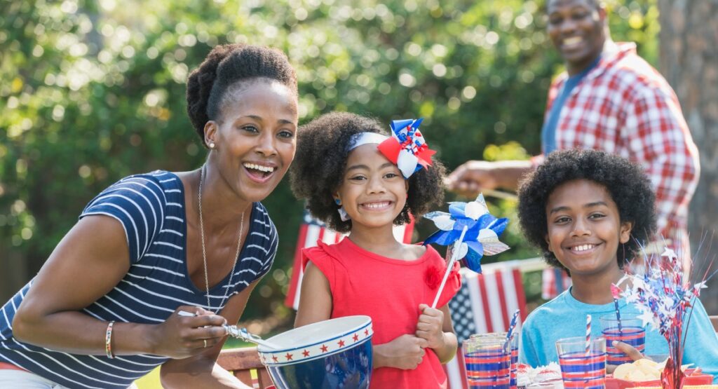 Family of 4 at an outdoor barbecue getting ready to celebrate July 4th. Family is wearing the colors red, white and blue. 
